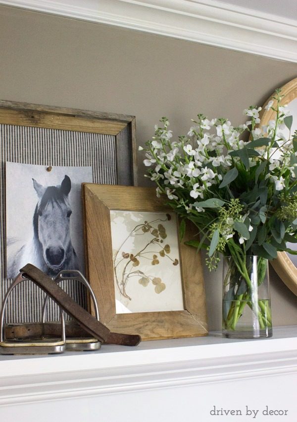 Fireplace mantel decorated with layered art, a round mirror, and flowers