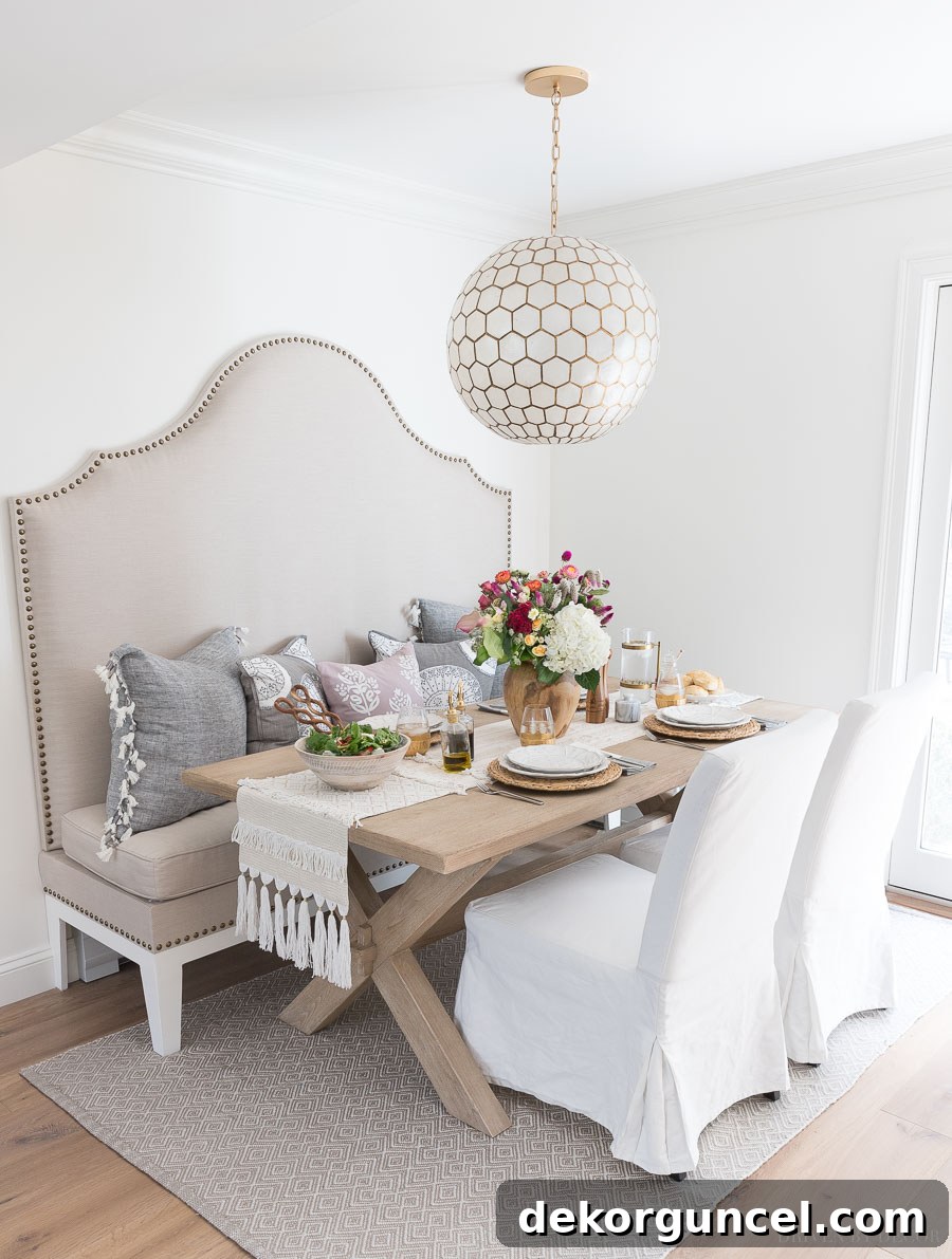The stunning transformation of the kitchen eat-in area, featuring a tall upholstered banquette, a warm wood trestle table, comfortable slipcovered dining chairs, and a captivating capiz pendant light.