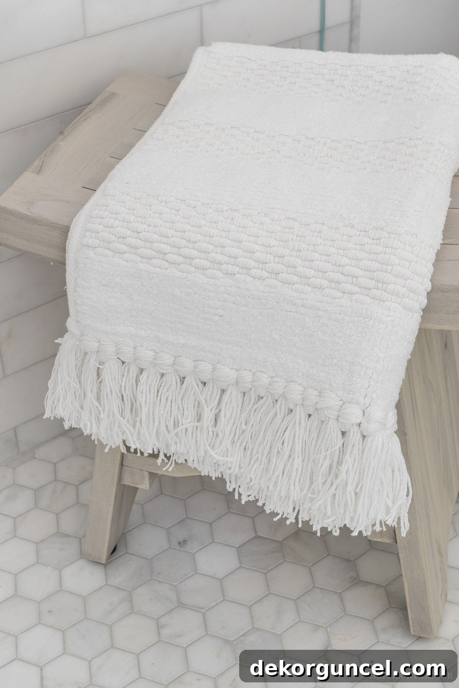 A charming tasseled bath mat resting on a small teak shower bench in the corner of the marble shower