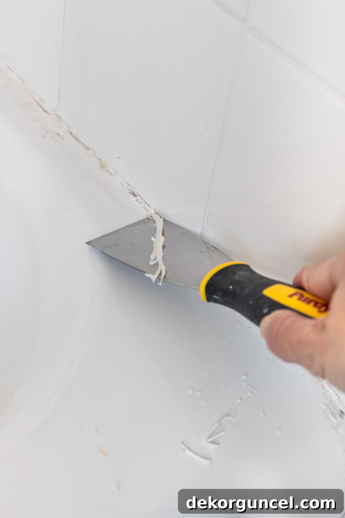 Using a stiff putty knife to scrape off the bottom edge of old caulk on a bathtub